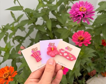 May include: Three clay pins on individual white cards labeled Bless Style. The pins feature a pink moka pot, a bright pink hibiscus flower, and a pink rectangular bar with white and gold flecks. The pins are held against a background of flowers.