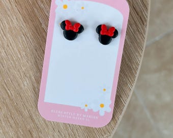 May include: A pair of black stud earrings shaped like mouse heads with red bows, displayed on a pink rectangular card featuring white daisy illustrations and the text Bless Style By Marian Winter Haven FL.