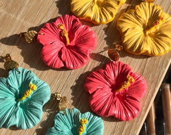 May include: Three pairs of hibiscus flower earrings in yellow, pink, and turquoise. Each earring features a textured five-petal flower with a prominent yellow stamen and a small, round, textured gold-tone metal stud attachment at the top.