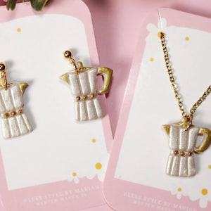 May include: A set of coffee-themed jewelry, including earrings and a necklace. The earrings and pendant feature a white and gold moka pot design. The jewelry is displayed on pink cards with the text "Bless Style by Marian Winter Haven FL".