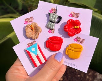 May include: A collection of five small decorative pins on white display cards labeled Bless Style. The pins feature a tan hibiscus, a silver moka pot, a red hibiscus, a Puerto Rican flag, a red mug, and a yellow mug.