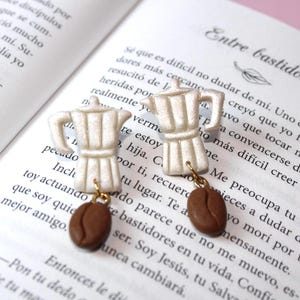May include: A pair of white ceramic earrings shaped like coffee pots with brown coffee bean charms hanging from them. The earrings are on a book with Spanish text.