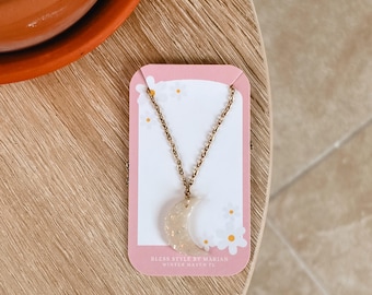 May include: A gold-toned chain necklace featuring a crescent moon pendant made of translucent material with embedded gold flecks. The necklace is displayed on a pink card with white daisy graphics and the text Bless Style By Marian Winter Haven FL.