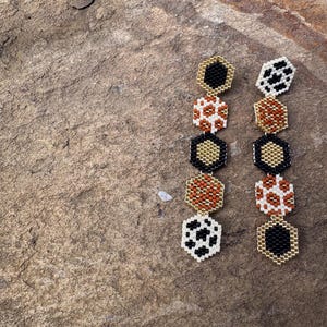 May include: A pair of long, dangling earrings with a geometric design. Each earring features five hexagonal beads with different animal print patterns in black, white, brown, and gold.