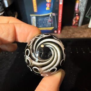 May include: A black and white glass marble with a swirling pattern and white circles. The marble has a clear center.