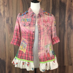 May include: A pink and blue plaid flannel shirt with a ruffle trim. The shirt has been decorated with comic book character patches, including Archie, Jughead, and Betty.