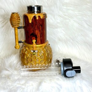 May include: A decorative water bottle with a honey-themed design. The bottle features a wooden-look body with gold accents, a honeycomb base, and a stainless steel lid. A wooden honey dipper and a small bee are also included.