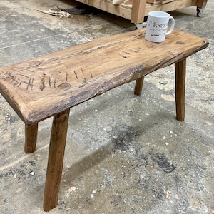 May include: A rustic wooden bench with a natural wood finish and three legs. The bench has a rough, textured surface and is made from reclaimed wood. The bench is sitting on a concrete floor.
