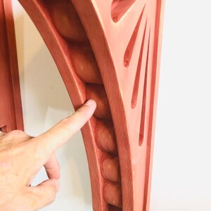 May include: A wooden shelf bracket with a unique design featuring a series of rounded protrusions along the curved edge and a sunburst pattern on the flat side. The bracket is painted a light pink color.