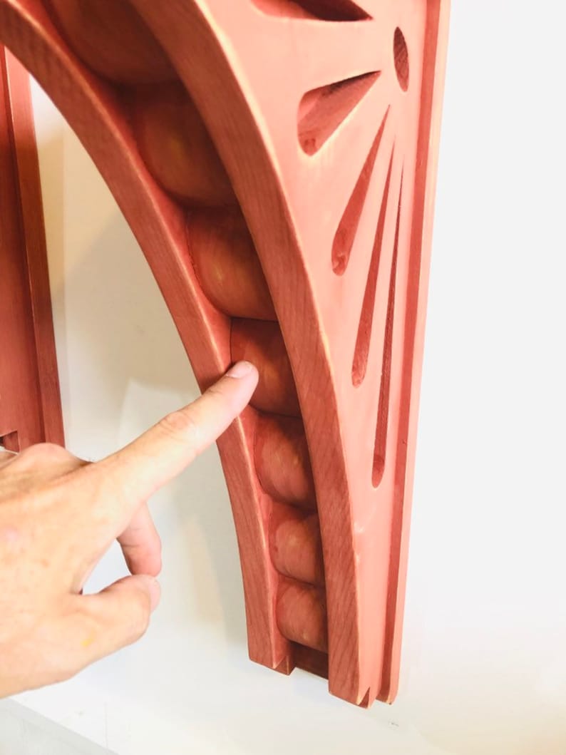 May include: A wooden shelf bracket with a unique design featuring a series of rounded protrusions along the curved edge and a sunburst pattern on the flat side. The bracket is painted a light pink color.