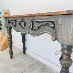 Peut inclure: Une console grise avec un plateau en bois sculpt&eacute; et des pieds tourn&eacute;s. La table a une finition vieillie et un aspect rustique.