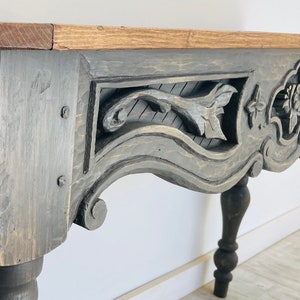 Peut inclure: Une table en bois gris avec des d&eacute;tails sculpt&eacute;s complexes. La table a un dessus incurv&eacute; et des pieds tourn&eacute;s. La sculpture pr&eacute;sente un motif floral avec des feuilles et des fleurs.