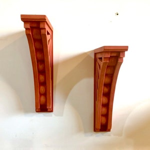 May include: Two wooden wall brackets with a decorative design. The brackets are painted a reddish-brown color and have a curved shape.