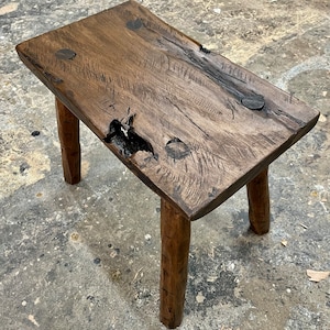 May include: A rustic wooden stool with a natural wood finish and four legs. The top of the stool is made of a single piece of wood with knots and grain patterns.