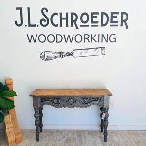 Peut inclure: Une console grise avec un plateau en bois sculpt&eacute; et des pieds tourn&eacute;s. La table est devant un mur blanc avec une enseigne noire et blanche qui indique "J.L. Schroeder Woodworking" et un dessin de ciseau.