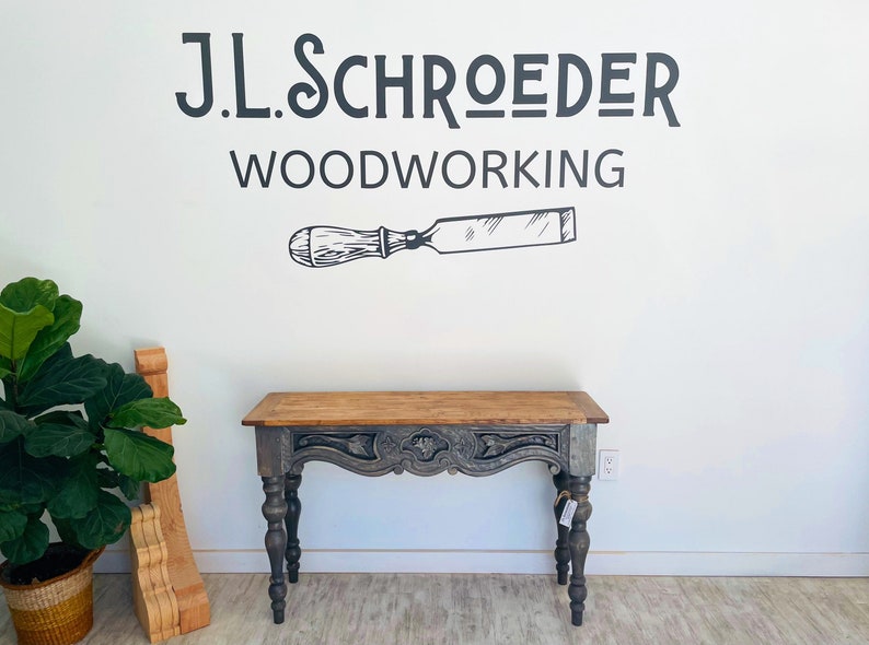 Peut inclure: Une console grise avec un plateau en bois sculpt&eacute; et des pieds tourn&eacute;s. La table est devant un mur blanc avec une enseigne noire et blanche qui indique "J.L. Schroeder Woodworking" et un dessin de ciseau.