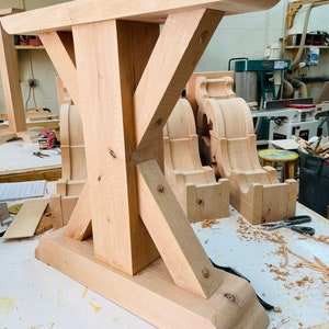 May include: A close-up of a wooden table base with a unique X-shaped design. The base is made of light-colored wood and has a smooth, polished finish. The base is sitting on a white surface with wood shavings scattered around it.