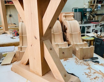 May include: A close-up of a wooden table base with a unique X-shaped design. The base is made of light-colored wood and has a smooth, polished finish. The base is sitting on a white surface with wood shavings scattered around it.