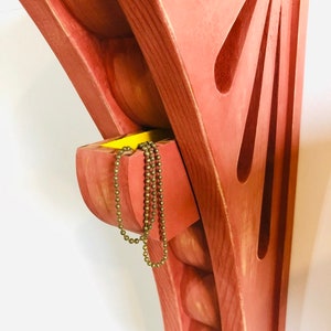 May include: A red wooden wall shelf with a small drawer. The drawer is open and a gold chain necklace is hanging out of it. The shelf has a unique design with rounded edges and a sunburst pattern.
