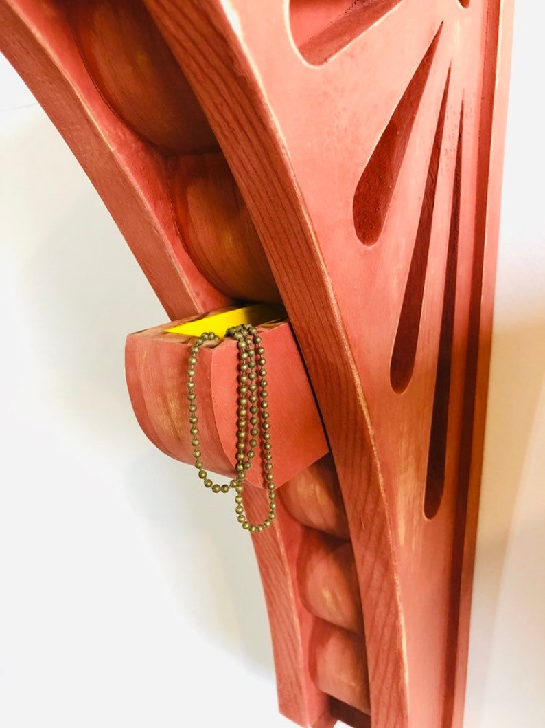 May include: A red wooden wall shelf with a small drawer. The drawer is open and a gold chain necklace is hanging out of it. The shelf has a unique design with rounded edges and a sunburst pattern.