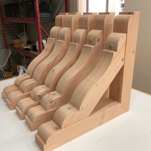May include: Five unfinished wooden corbels, each with a decorative scroll design, are arranged in a row on a light wood surface. The corbels are made of light-colored wood and have a natural finish.