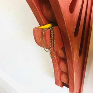 May include: A red wooden wall shelf with a small yellow drawer and a chain hanging out of it. The shelf has a decorative design with rounded edges and a fan-shaped cutout.