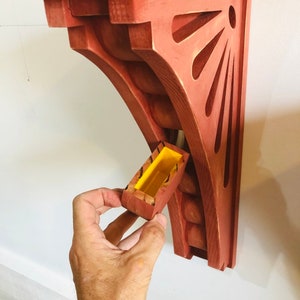 May include: A red wooden shelf bracket with a decorative cutout design and a small yellow box attached to the side.