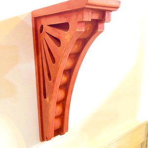 May include: A red wooden shelf bracket with a decorative sunburst design and a rounded edge. The bracket is made of solid wood and has a distressed finish.
