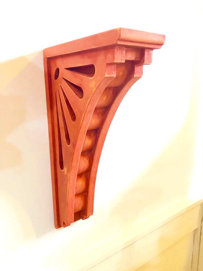 May include: A red wooden shelf bracket with a decorative sunburst design and a rounded edge. The bracket is made of solid wood and has a distressed finish.