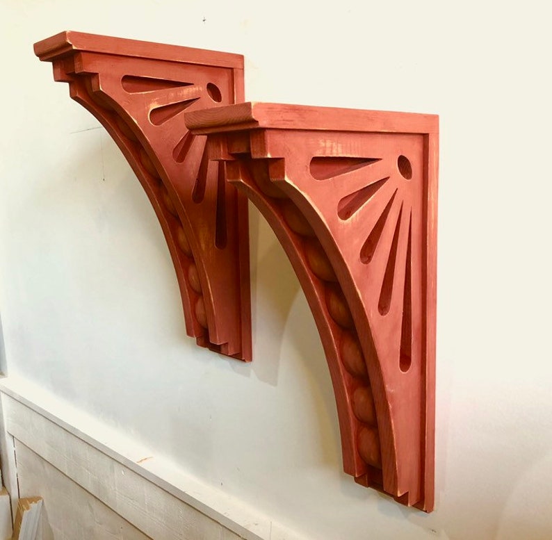 May include: Two red wooden corbel brackets with a decorative sunburst design and a beaded edge. The brackets are mounted on a white wall.