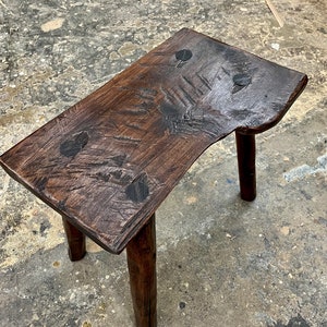 May include: A rustic wooden stool with a dark brown finish and four legs. The top of the stool is uneven and has a unique, hand-crafted look.