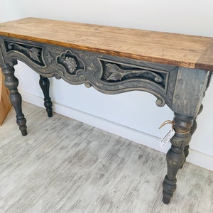 Peut inclure: Une console grise avec un motif en bois sculpt&eacute; et un plateau en bois naturel. La table a des pieds tourn&eacute;s et une finition vieillie.