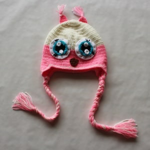 May include: A pink and white crocheted owl hat with long pink and white braided ties. The hat has large blue eyes with white and black accents.