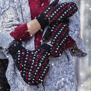 May include: A pair of black mittens with red and white hearts. The mittens are worn by a person wearing a grey sweater and blue jeans.