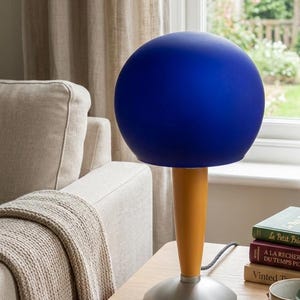 May include: A modern table lamp with a spherical, cobalt blue glass shade. The lamp has a tapered, yellow-orange stem and a circular, silver-colored base. The lamp is on a wooden table next to a sofa and books.