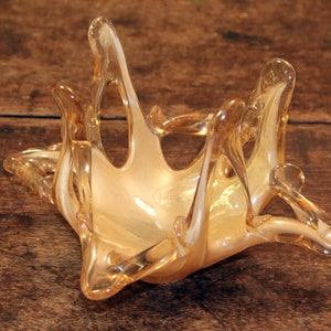 May include: A clear glass bowl with a unique, abstract design. The bowl is made of blown glass and has a light amber color with a white iridescent finish.
