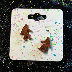 May include: A pair of brown glitter Bigfoot earrings on a white card with colorful splatter paint.