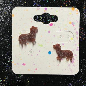 May include: A pair of brown dachshund-shaped earrings with glitter on a white card with colorful splatter paint.