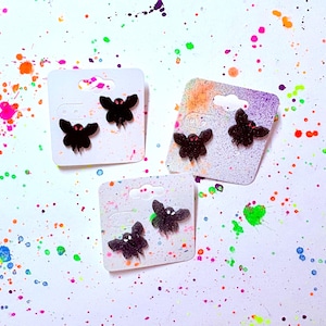 May include: Three pairs of black bat-shaped earrings on white earring cards. The earrings have red accents. The background is white with colorful paint splatters in pink, orange, green, blue, and purple.