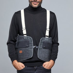 May include: A man wearing a black turtleneck sweater with a denim vest featuring white straps and multiple pockets. The vest has a black patch with a logo on the front.