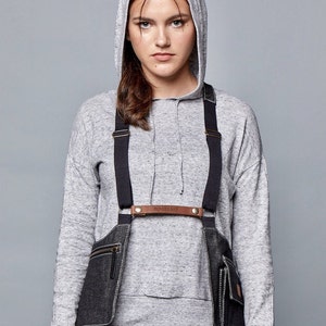 以下が含まれることがあります： 黒いストラップと真鍮色の金具が付いたデニムのチェストハーネス。 ハーネスには、ジッパー付きのフロントポケットが2つと、胸に「MOVING BAGS」の文字が入った革バンドが付いています。 モデルはグレーのフード付きスウェットシャツを着ています。