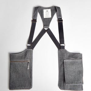 May include: Gray denim apron with two front pockets, one with a zipper, and a smaller pocket. The apron has black straps and white stitching. A tag with text is visible at the top.