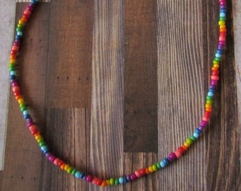 May include: A rainbow-colored beaded necklace with a silver-tone chain and clasp. The necklace features small, round beads in a spectrum of colors, including red, orange, yellow, green, blue, and purple. The necklace is displayed against a wooden background.