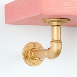 Könnte beinhalten: Ein rosa Holzregal mit goldfarbenen Rohrbügeln. Das Regal ist an einer weißen Wand befestigt. Die Halterungen haben eine kreisförmige Basis mit dem Text "DS 1914".