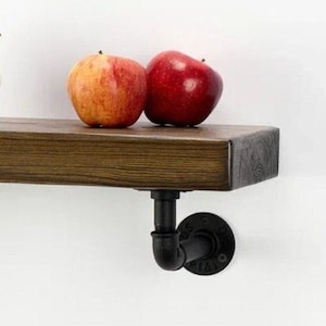 Könnte beinhalten: Ein rustikales Holzregal mit dunkler Oberfläche, getragen von einem schwarzen Rohrfitting. Zwei Äpfel, einer rot und einer orange, liegen auf dem Regal. Das Regal ist an einer weißen Wand befestigt.