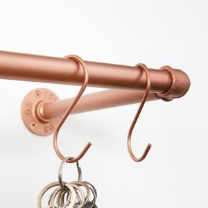 Pode incluir: Uma prateleira de tubo de cobre com dois ganchos presos. Os ganchos são de metal e têm a forma de um 'J'. Um conjunto de chaves está pendurado em um dos ganchos.