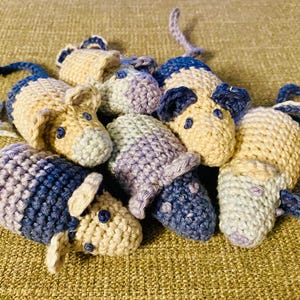 May include: A collection of handmade crochet mice in various colors. The mice are primarily beige, blue, and purple, with small black eyes and long tails. The toys are clustered together on a textured surface.