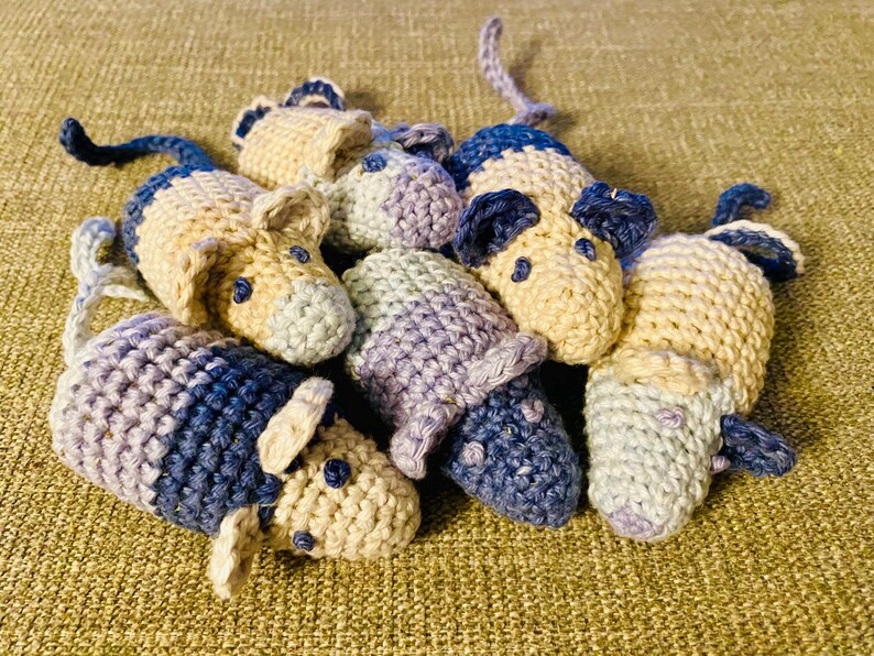 May include: A collection of handmade crochet mice in various colors. The mice are primarily beige, blue, and purple, with small black eyes and long tails. The toys are clustered together on a textured surface.