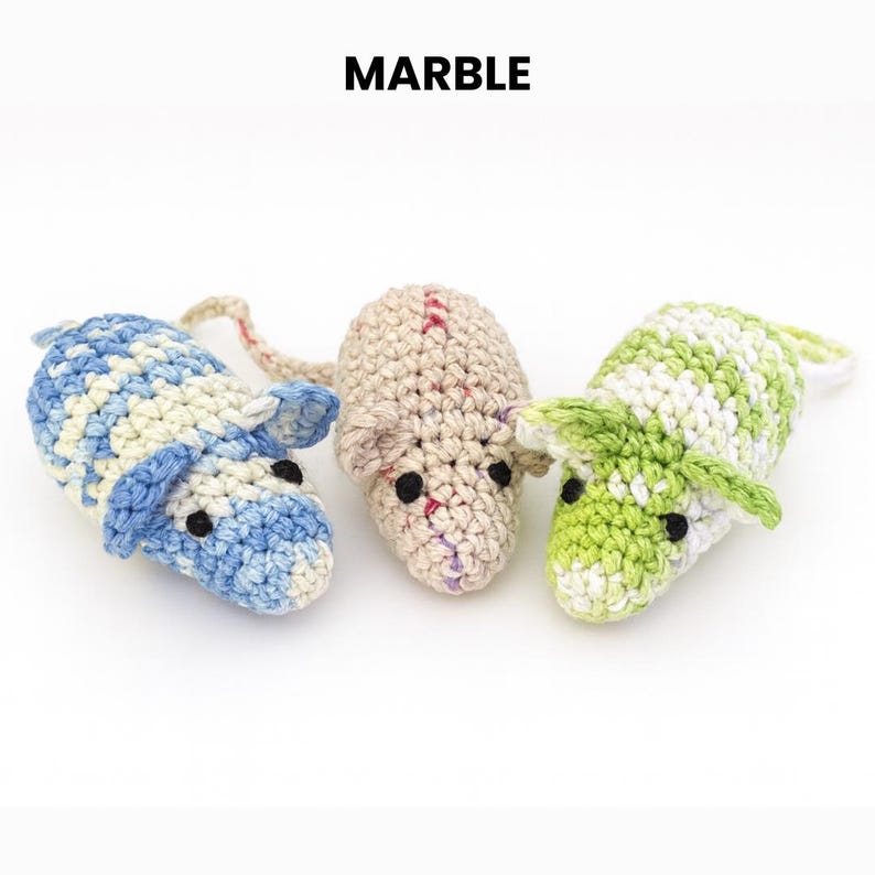 May include: Three crocheted mouse toys in blue and white, tan, and green and white. The word "MARBLE" is printed in black above the toys. Each mouse has black eyes and a long tail.