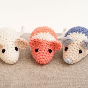 May include: Three handmade crochet mice in a row. One is off-white, one is coral with white stripes, and the last is blue and beige. Each mouse has black eyes, beige ears, and a tail.